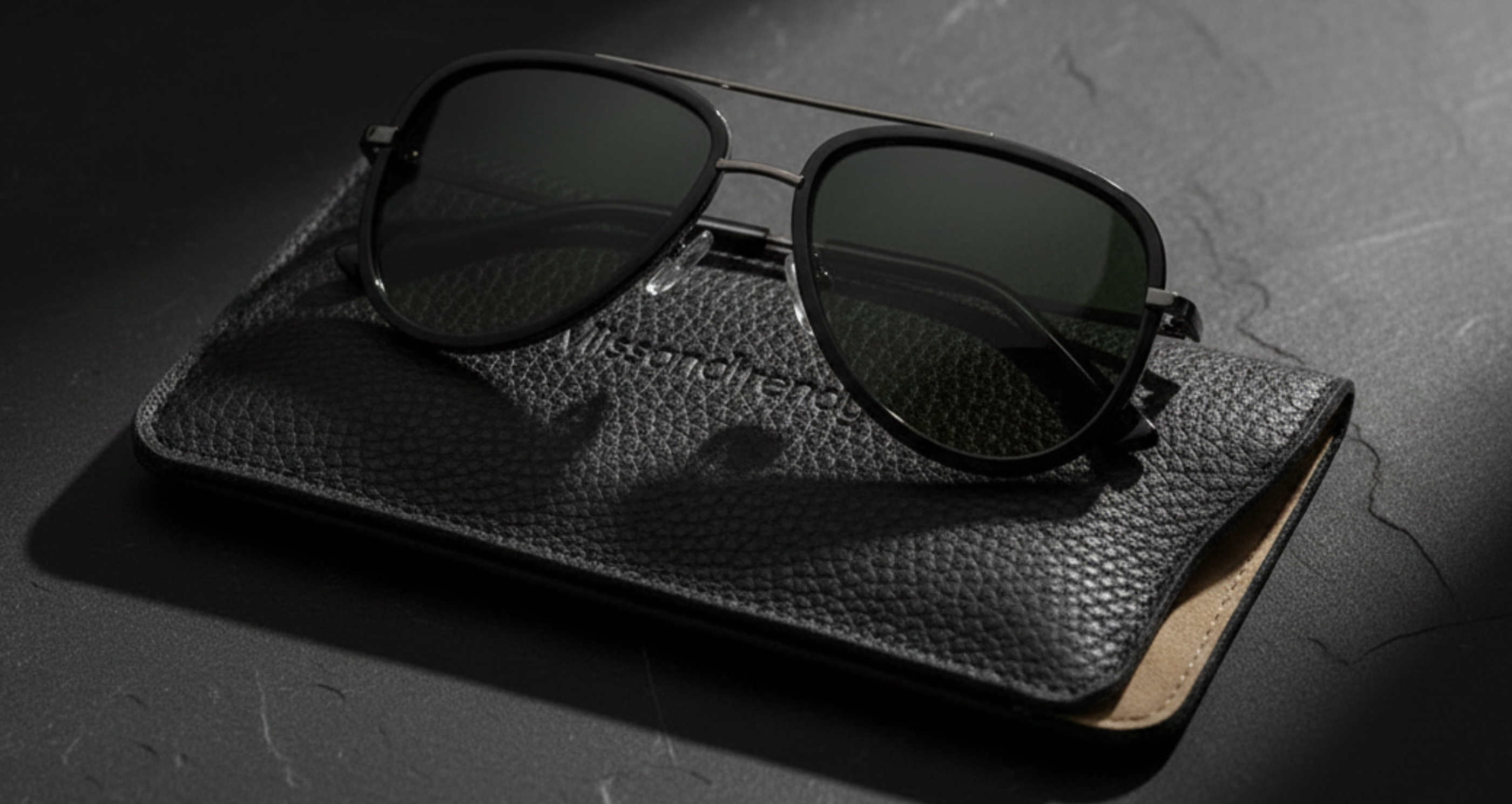 Dallas - Black aviator sunglasses with dark lenses resting on a textured black leather case, set against a dark gray stone background - Missandtrendy
