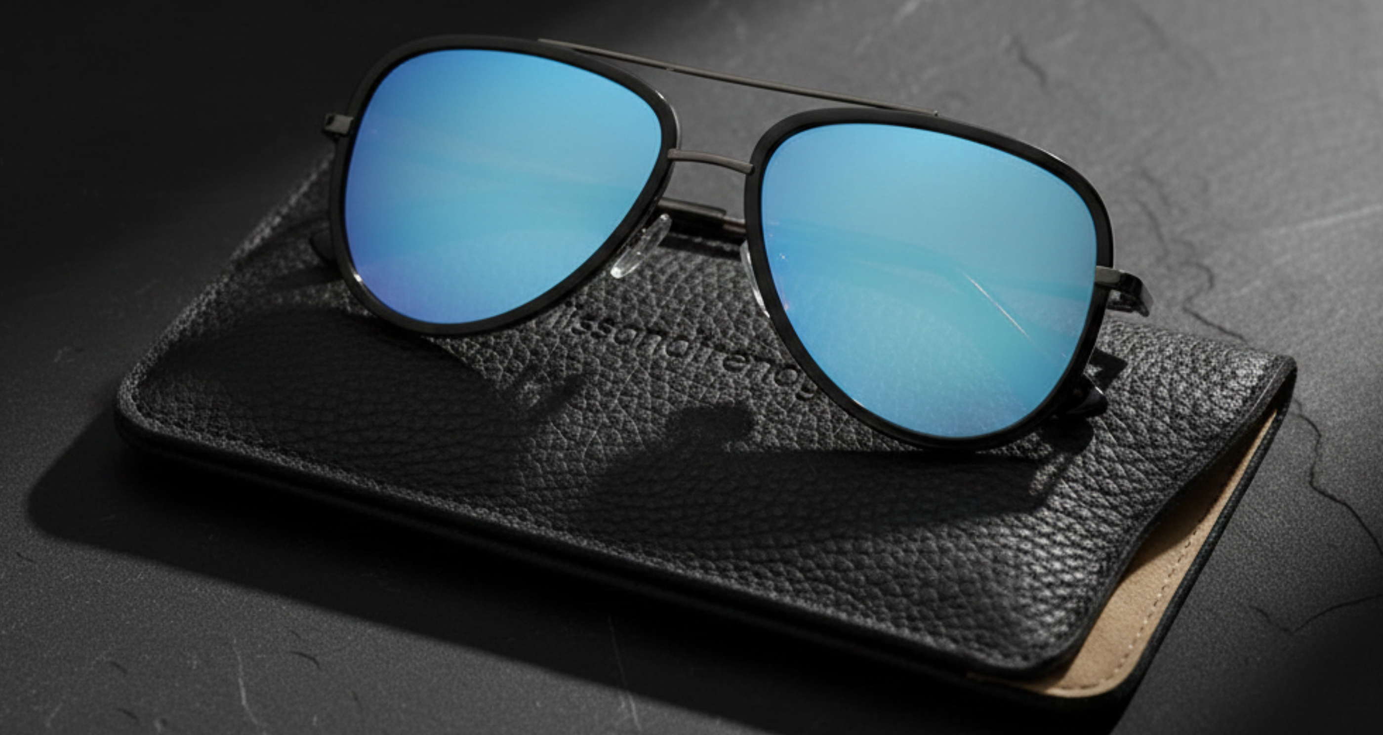 Dallas Mirror - A pair of aviator-style sunglasses with blue mirrored UV 400 polarised lenses and black frames, resting on a textured black leather case against a dark gray background _ Missandtrendy