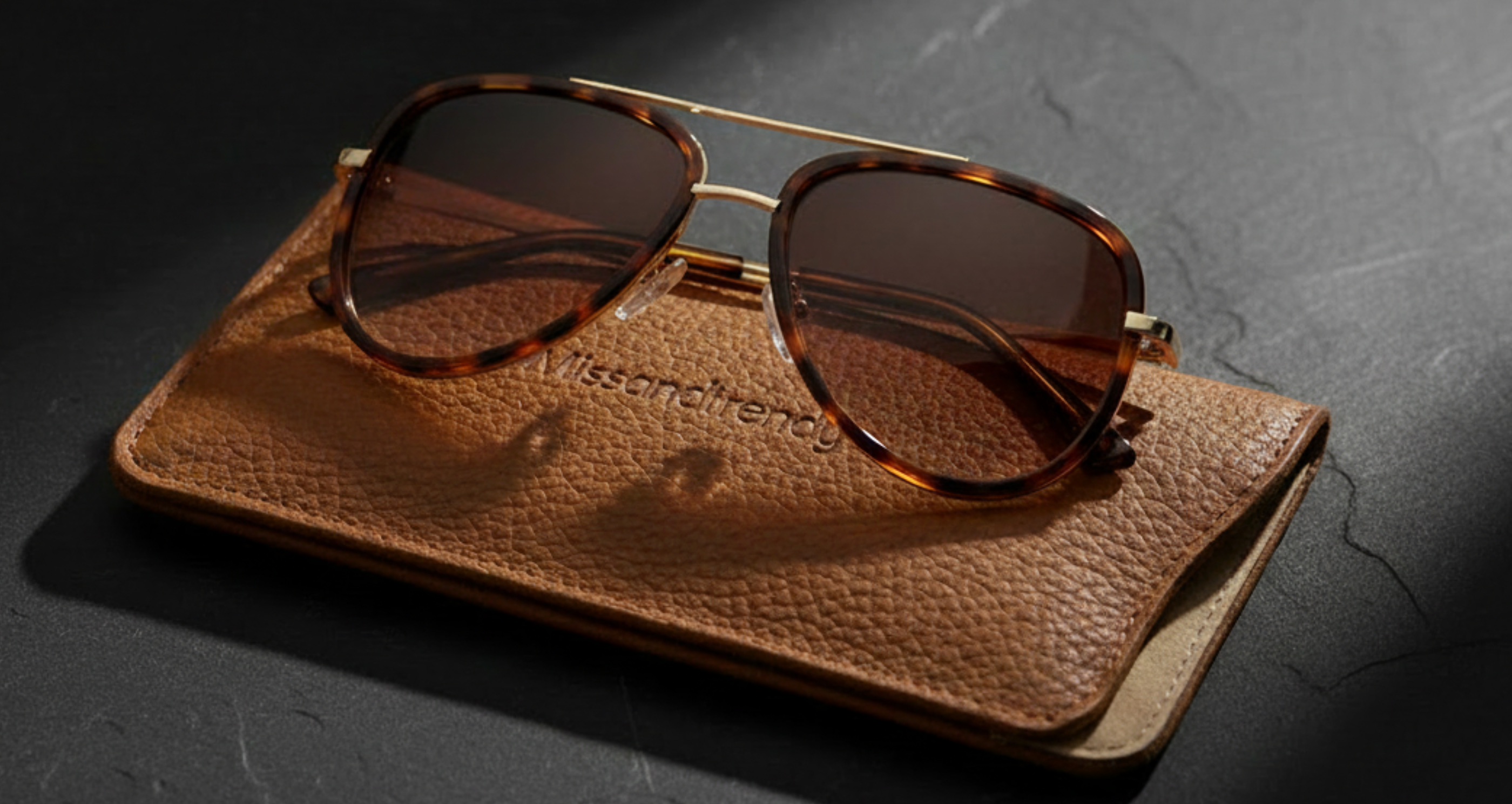 Dallas - A pair of aviator-style sunglasses with tortoiseshell frames and dark lenses resting on a brown textured leather case, photographed on a dark stone surface with dramatic side lighting - Missandtrendy