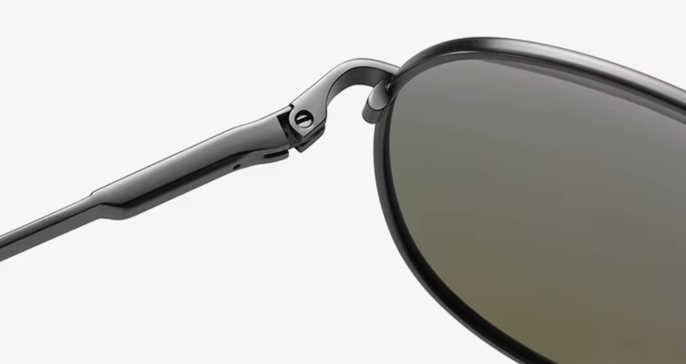 Close-up of a pair of sunglasses showing the dark gray temple arm, hinge, and part of the dark gray lens against a light gray background.