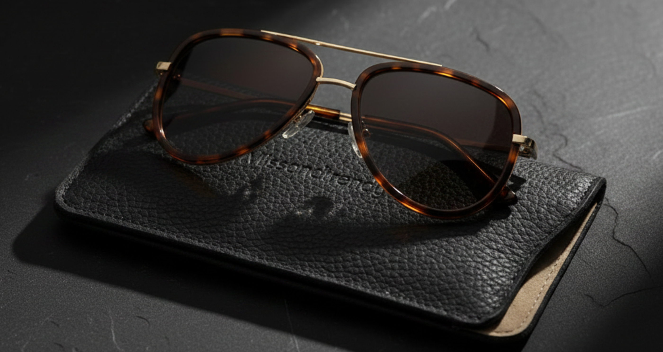 Dallas - A pair of aviator-style sunglasses with tortoiseshell frames and dark lenses, resting on a black textured leather case against a dark stone background - Missandtrendy