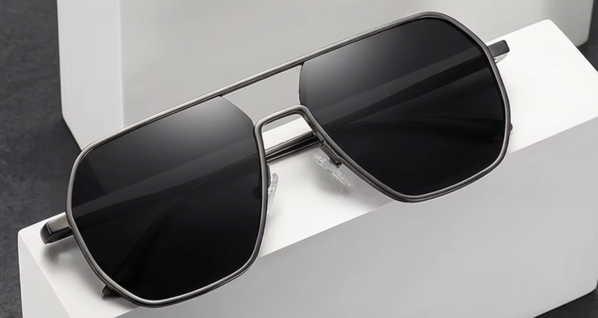 A pair of angular aviator-style photochomic UV polarised sunglasses with a dark gray metal frame and black lenses, resting on a white display block.