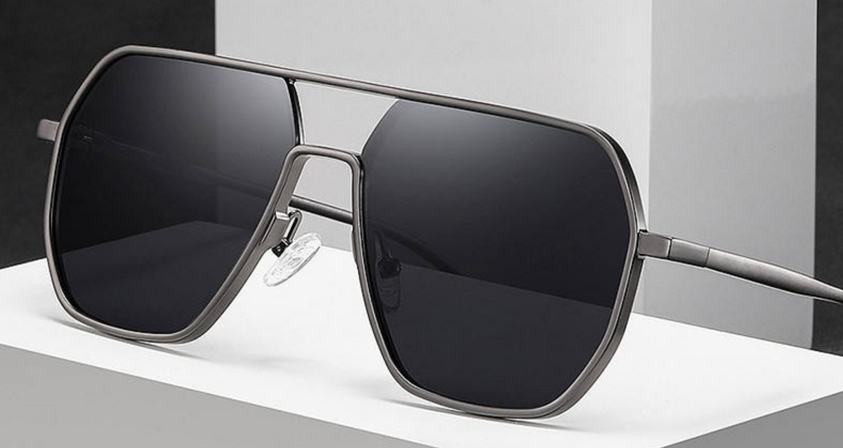 A pair of oversized aviator-style photochomic UV polarised sunglasses with a matte dark gray metal frame and dark tinted lenses, displayed on a white geometric platform.