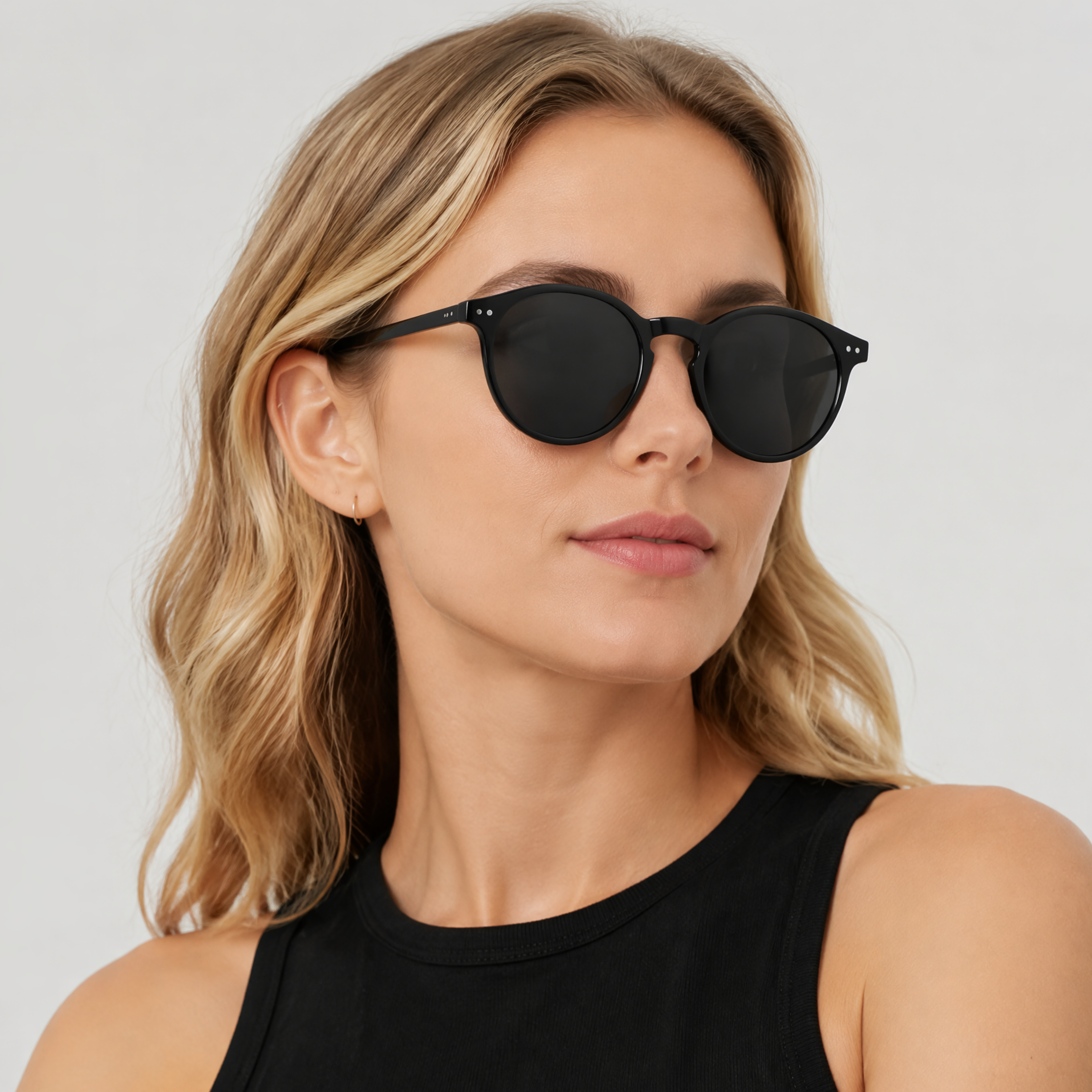 Sevilla - A close-up of a woman with blonde hair wearing black round sunglasses and a black sleeveless top, photographed against a light gray studio background - Missandtrendy